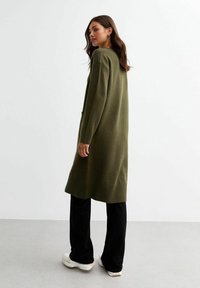 Cardigan lungo verde oliva realizzato in materiale a maglia testurizzato, con vestibilità ampia e orli a coste, abbinato a pantaloni neri a gamba larga e scarpe bianche.