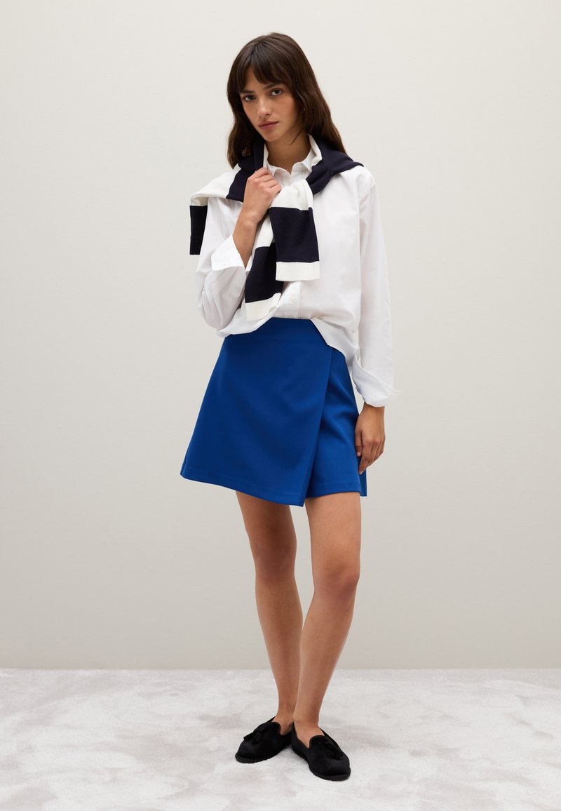 Camisa blanca de botones, falda envolvente azul, zapatos de lona negros. Suéter azul marino drapeado sobre los hombros. Tejido suave, corte relajado y líneas limpias.