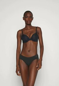 Feles áll egy egyszínű háttér előtt, fekete melltartót és hozzá illő fekete alsónemût viselve, szembenézve a kamerával, arckifejezése semleges.