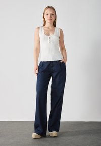 White ribbed tank top with scalloped edges and three buttons, paired with dark blue wide-leg denim pants. Model stands with hands in pockets.