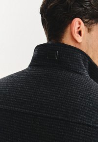 Czarny wełniany płaszcz teksturowany z wysokim kołnierzem, z prostokątną metką marki z tyłu. Zawiera zbliżenie na kołnierz i detal tkaniny.