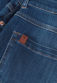 Jeans de mezclilla azul oscuro con costuras naranjas contrastantes, que presentan una pequeña etiqueta de cuero marrón en el bolsillo trasero. La textura parece suave.