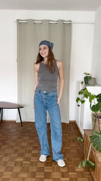 Top de tanque cinza com riscas, jeans claros azuis largos, ténis brancos; o fundo apresenta uma cortina verde e uma planta em vaso. O chão tem um padrão de madeira.
