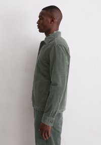 Zielona kurtka ze sztruksu z kołnierzem, pionowymi paskami, zapinanymi na guziki mankietami i luźnym krojem, pokazana na modelce z profilu na jednolitym tle.