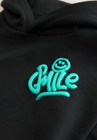 Sort hoodie med et teal broderet "Smile" logo med et smilende ansigt, hvilket skaber en tekstureret kontrast til stoffet.