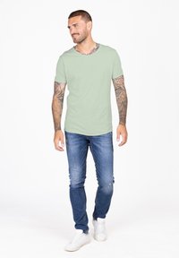 Camiseta de manga corta verde claro, combinada con jeans ajustados azules y zapatillas blancas. La tela parece suave, con una textura lisa.