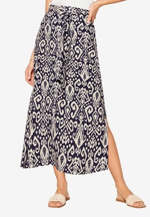 Femme portant une jupe midi noire et blanche à motifs avec une ceinture nouée à la taille et une fente latérale, associée à des sandales beige à bout ouvert.