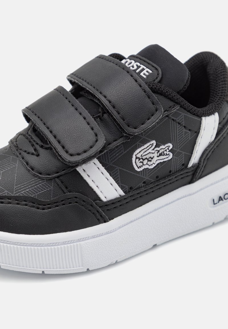 Czarne syntetyczne sneakersy z białymi akcentami, z geometrycznym wzorem, rzepami i teksturowaną białą gumową podeszwą. Wyświetlone logo Lacoste.