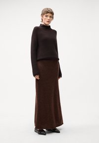 Maglione a collo alto in maglia marrone abbinato a una lunga gonna marrone uniforme. La gonna presenta una consistenza morbida, e l'outfit è completato da scarpe flat nere decorate con borchie metalliche.