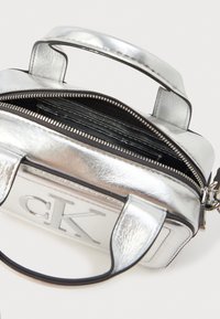 Srebrna metaliczna torebka z dwiema krótkimi rączkami, zamykana na zamek, z wytłoczonym logo. Ma fakturowaną powierzchnię i czarne kieszenie wewnętrzne.