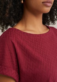 Burgundia tekstuuriga topp, millel on herringbone muster, lai kaelus ja lühikesed varrukad, valmistatud kergest ja hingavast kangast.