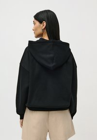 Woman with long black hair wearing silver hoop earrings, a black hooded sweatshirt, and beige shorts, facing away from viewer against plain background.