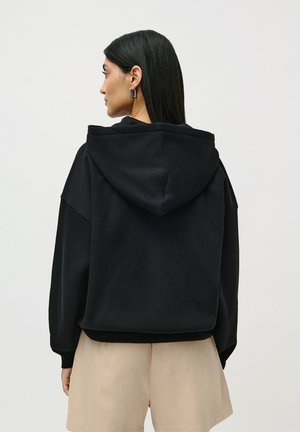 Kvinne med langt svart hår som har på seg sølvfargede hoops, en svart hettegenser og beige shortser, vendt bort fra betrakteren mot en ensfarget bakgrunn.