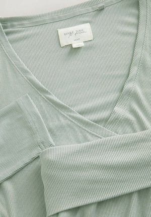 Tejido texturizado de color verde claro con un diseño de cuello en V y costuras visibles. La etiqueta dice "HAZ TIEMPO PARA SOÑAR" de la marca "NEXT."