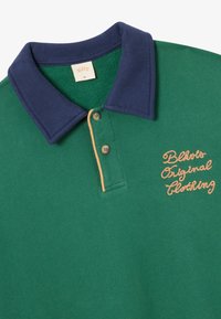 Zielona koszulka polo z granatowym kołnierzykiem, z pomarańczowym haftowanym logo, na którym widnieje napis "Blots Original Clothing" oraz z dwoma guzikami.