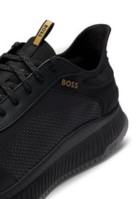 Baskets noires en tissu texturé, avec des accents en cuir lisse et une semelle crantée. Présentent un logo doré "BOSS" et des détails en motif sur le côté.