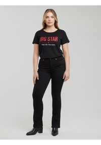 Nő áll, fekete "BIG STAR Your Life. Your Jeans." feliratú pólóban, fekete testhez álló farmerben és fekete, magas sarkú bokacsizmában, szürke háttér előtt.