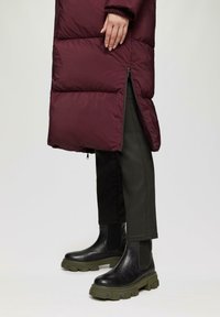Bordeauxkleurige pufferjas met verticale bafflepatroon en zijrits, gecombineerd met zwarte enkellaarsjes met een dikke groene zool en elastische zijpanelen.
