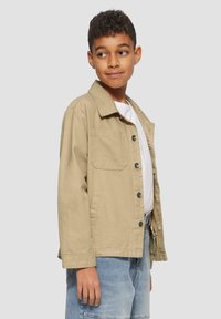 Khaki jas met een kraag, een voorzak en groene knopen, gedragen over een wit shirt. Het materiaal lijkt lichtgewicht en gestructureerd te zijn.