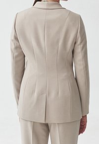 Blazer beige sur mesure avec une coupe structurée, doté d'épaules marquées, de pinces dans le dos et d'une fente unique, confectionné dans un tissu lisse.
