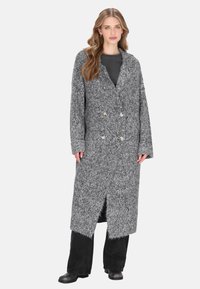 Donna che indossa un lungo cardigan grigio a maglia spessa con doppi bottoni sopra pantaloni neri e stivali, in piedi su uno sfondo bianco.