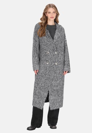 Vrouw in een lange, robuuste grijze gebreide cardigan met dubbele knopen, over zwarte broeken en laarzen, staande tegen een witte achtergrond.
