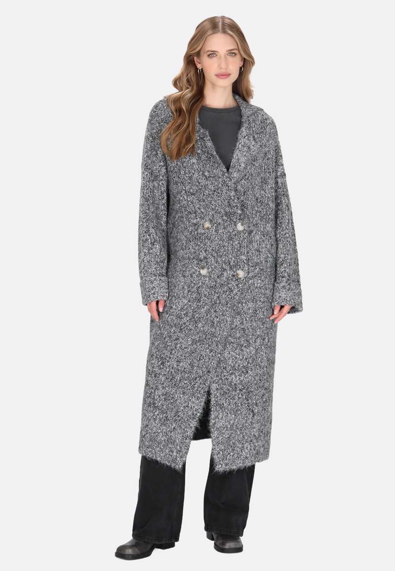 Donna che indossa un lungo cardigan grigio a maglia spessa con doppi bottoni sopra pantaloni neri e stivali, in piedi su uno sfondo bianco.
