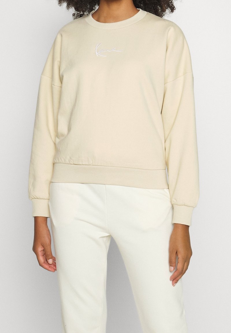 Femme portant un sweat-shirt beige à manches longues avec poignets retournés et un pantalon clair assorti sur un fond uni.