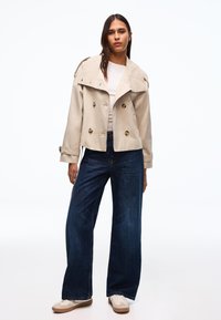 Frau steht und trägt einen beigen, gekürzten Trenchcoat, ein weißes Oberteil, weite dunkelblaue Jeans und weiße Turnschuhe vor weißem Hintergrund.