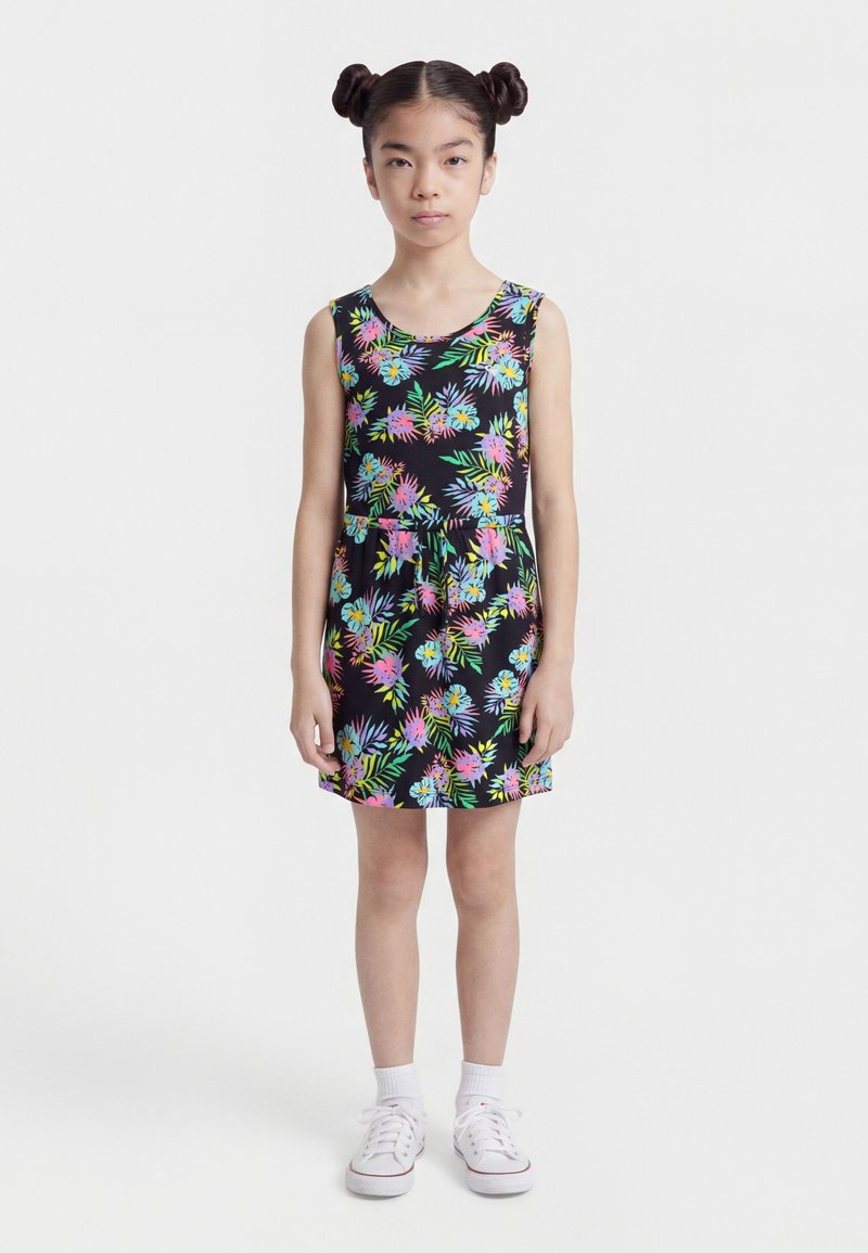 Νεαρό κορίτσι με διπλούς κότσους, φοράει αμάνικο μαύρο φόρεμα με πολύχρωμο λουλουδάτο σχέδιο και λευκά αθλητικά παπούτσια, στέκεται με το πρόσωπο μπροστά.