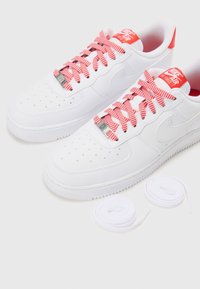 Baskets Nike Air Force 1 blanches avec des lacets à motif chevron rouge et blanc, des étiquettes de logo rouges et deux paires de lacets blancs supplémentaires placées à côté.