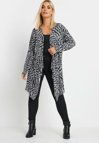 Cardigan leggero e aperto davanti di colore grigio con un motivo astratto nero. Presenta maniche lunghe e arriva sotto i fianchi, abbinato a pantaloni neri e stivali.