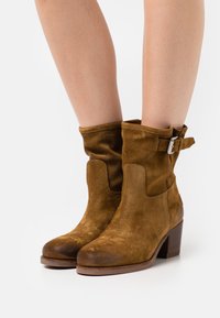 Botas de tobillo de ante marrón con un diseño arrugado, que cuentan con una hebilla lateral y un tacón grueso. Textura suave y punta redondeada.