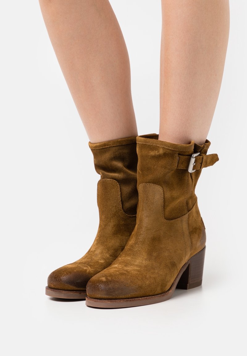 Botas de tobillo de ante marrón con un diseño arrugado, que cuentan con una hebilla lateral y un tacón grueso. Textura suave y punta redondeada.