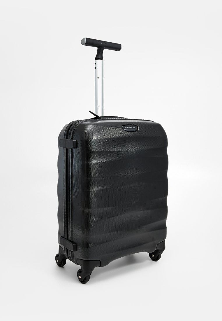 Valise rigide noire avec surface texturée, poignée télescopique et quatre roues. Présente une fermeture éclair et un accent de marque.