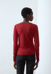Long-sleeve red top in stretch fabric with a round neckline and ruched detailing along the sleeves, paired with dark jeans.