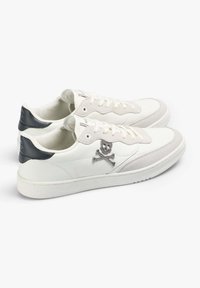 Adidași din piele albă cu accente gri, textură netedă, talpă plată și detalii brodate cu craniul și oasele încrucișate pe lateral.