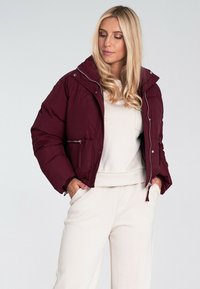 Bordowa kurtka puchowa z wysokim kołnierzem, zamkiem z przodu i bocznymi kieszeniami. Gładka faktura z pikowanym wzorem; noszona na lekkiej bluzce.