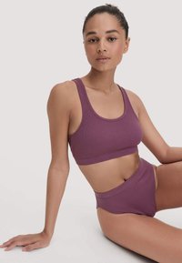 Jonge vrouw zittend op de vloer in een bijpassende ribbelige paarse sport-bh en high-waisted ondergoed tegen een effen lichte achtergrond.