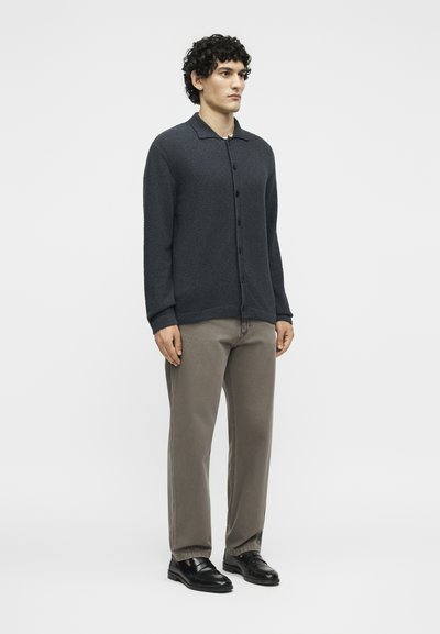 Άνδρας με σγουρά μαλλιά που φοράει σκούρη ζακέτα με κουμπιά, γκρι παντελόνι και μαύρα δερμάτινα παπούτσια, στέκεται μπροστά σε ένα απλό ανοιχτό φόντο.