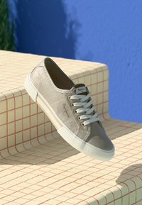 Baskets en toile beige avec des lacets blancs, surface texturée et semelle en caoutchouc. Présente un branding discret et se trouve sur une surface à motif écossais.