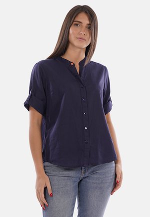 Femme aux cheveux bruns lisses portant une chemise bleu marine courte à manches courtes avec boutons et un jean bleu clair, debout devant un fond uni.