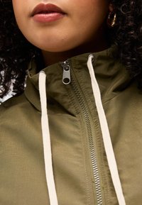 Κοντινό πλάνο του κάτω μέρους προσώπου ενός ατόμου που φοράει ένα χακί πράσινο μπουφάν με λευκά κορδόνια και ασημένια φερμουάρ που είναι μερικώς ανοιγμένο.