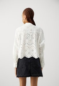 Blusa bianca in pizzo con orlo scallop e maniche lunghe, che mostra intricati motivi ritagliati. Abbinata a una mini gonna in denim scuro.