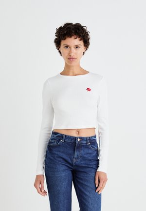 White long-sleeve cropped top with ribbed texture and a small red rose embroidery on the left chest, paired with dark blue high-waisted jeans.