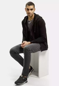 Czarna rozpinana bluza na suwak na szaro, z szarą koszulką, szarymi spodniami typu slim-fit i czarnymi trampkami. Model siedzi na białym bloku.