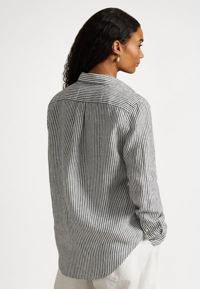 Γυναίκα με σκούρα σγουρά μαλλιά, που φοράει μαύρο-άσπρο πουκάμισο με κάθετες ρίγες μακριάς μανσέτας και ανοιχτόχρωμο παντελόνι, φαίνεται από την πλάτη.