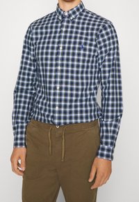 Modrá a zelená kostkovaná košile s knoflíky, bílými akcenty a límečkem, nošena s béžovými kalhotami na stažení.