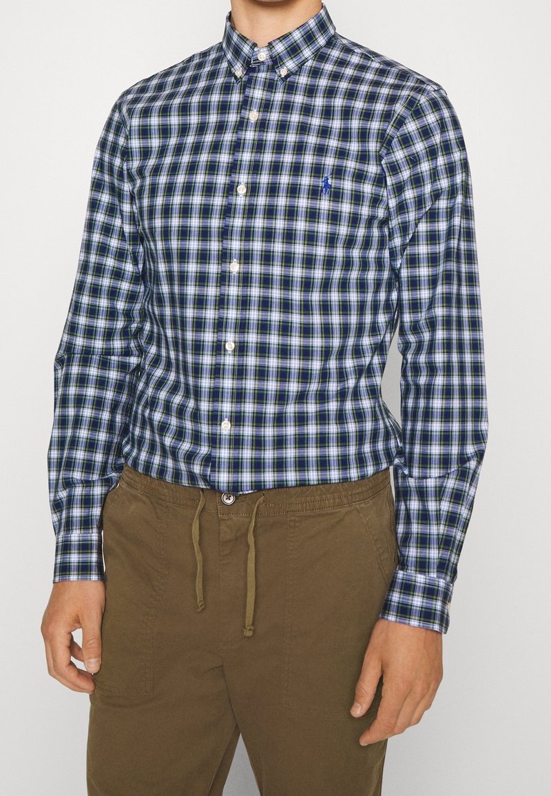 Modrá a zelená kostkovaná košile s knoflíky, bílými akcenty a límečkem, nošena s béžovými kalhotami na stažení.