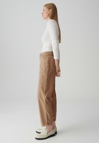 Pantalons en velours côtelé beige avec des jambes larges et des poches latérales proéminentes, associés à un haut ajusté à manches longues blanc et des mocassins de couleur claire.
